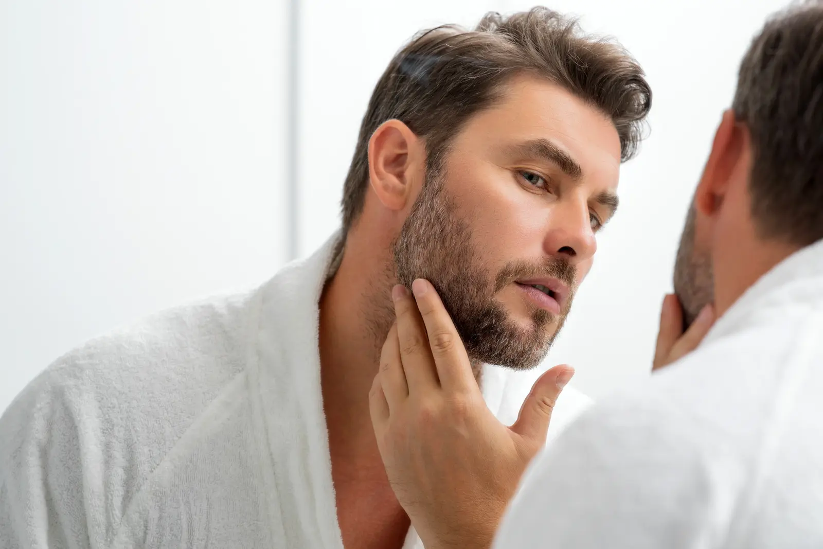 Man examining his face in the mirror, wearing a white robe.