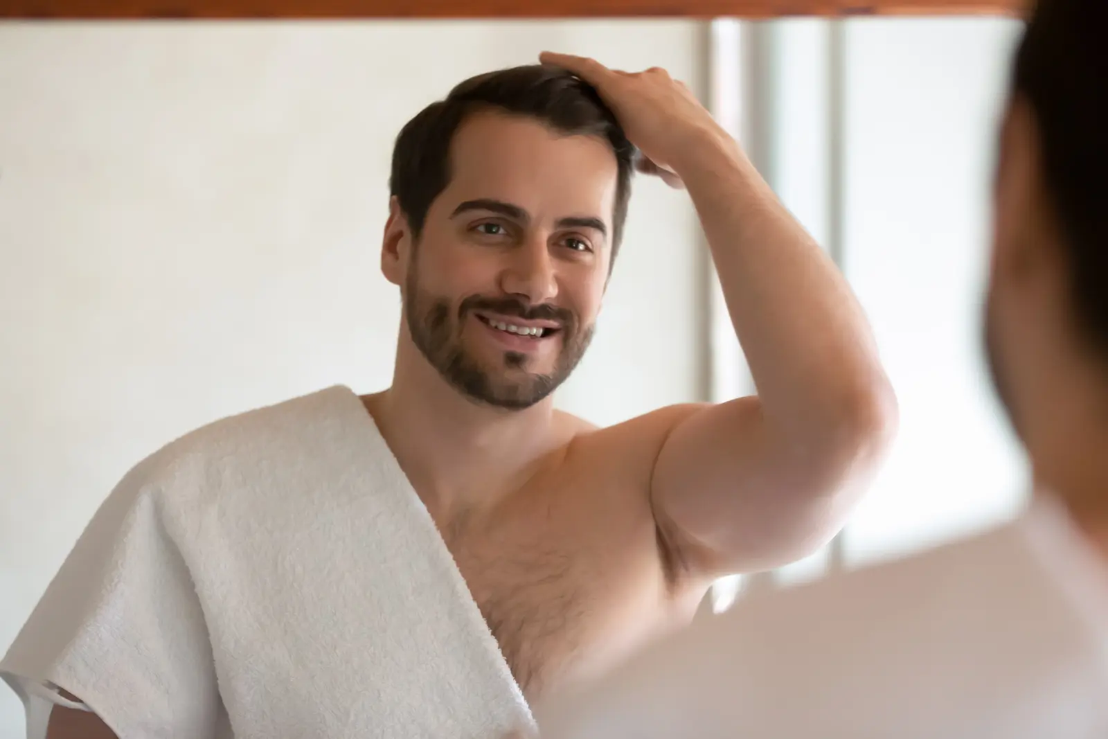 Smiling man with towel looking in mirror, touching hair.